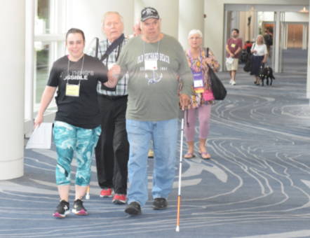 A group of people are walking through a spacious hallway. In front are a man with a white cane and a volunteer guiding him.