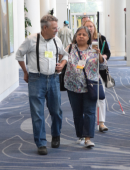 A volunteer is walking with a woman and they appear engaged in conversation, The are walking through a hallway at a convention.